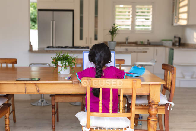 Chica hispana sentada en la mesa de la cocina haciendo trabajo escolar usando laptop. familia feliz en casa. - foto de stock