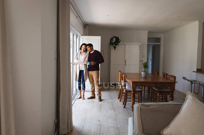 Couple hispanique debout dans la cuisine buvant du café et regardant par la fenêtre. passer du temps ensemble à la maison. — Photo de stock