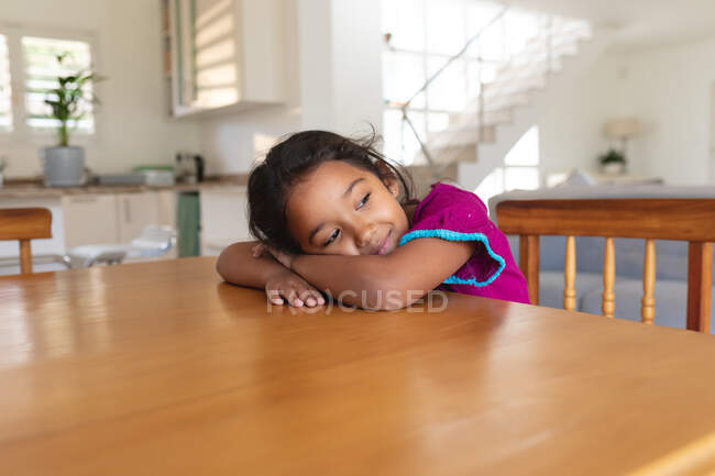 Sorridente ragazza ispanica seduta a tavola in cucina poggiando la testa sulle braccia, distogliendo lo sguardo. trascorrere del tempo libero a casa. — Foto stock