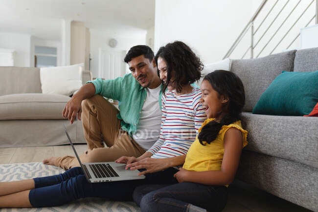 Sonriendo madre hispana, padre e hija sentados en el piso de la sala de estar usando un portátil juntos. familia pasar tiempo juntos en casa. - foto de stock