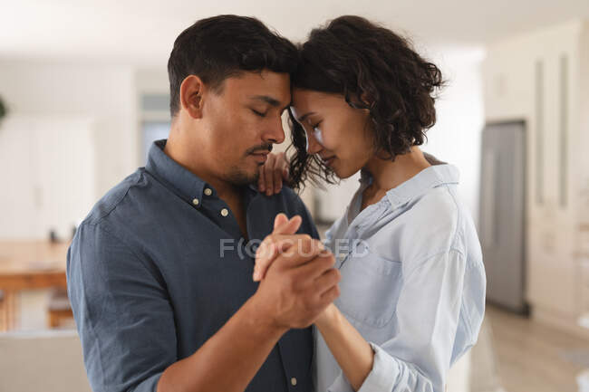 Romántica pareja hispana bailando lentamente en la sala de estar con los ojos cerrados. en casa en aislamiento durante el bloqueo de cuarentena. - foto de stock