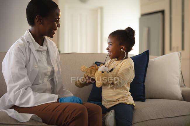 Una doctora afroamericana sonriente visitando a una paciente en casa, jugando con un estetoscopio. servicios médicos y sanitarios, visita al médico en el hogar. - foto de stock