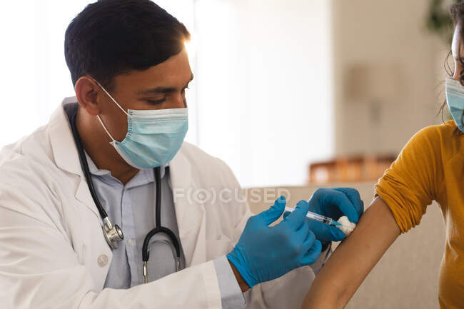 Médico masculino hispano que da la vacuna covid al paciente femenino en casa, usando máscaras faciales. servicios médicos y sanitarios durante la pandemia de coronavirus covid 19. - foto de stock