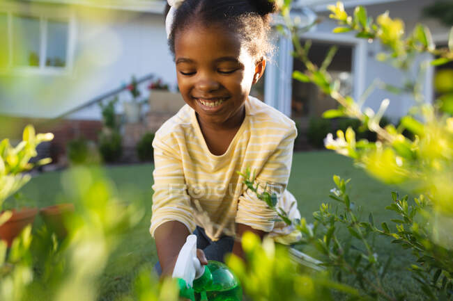 Lächelnde afrikanische Amerikanerin beim Gärtnern, Knien und Gießen von Pflanzen im sonnigen Garten. Freizeit zu Hause verbringen. — Stockfoto