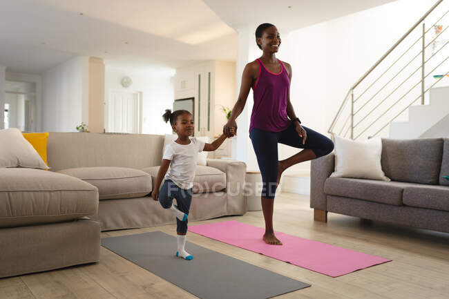 Sonriendo afroamericana madre e hija practicando yoga cogidas de la mano y de pie sobre una pierna. familia pasar tiempo juntos en casa. - foto de stock