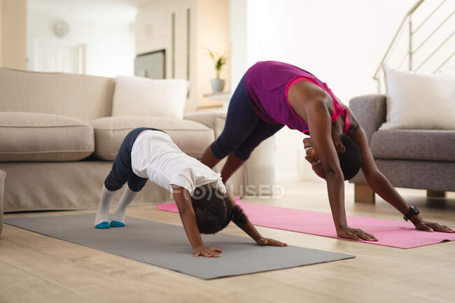 Mère et fille afro-américaine souriante pratiquant le yoga, s'étirant sur des nattes dans le salon. passer du temps ensemble à la maison. — Photo de stock