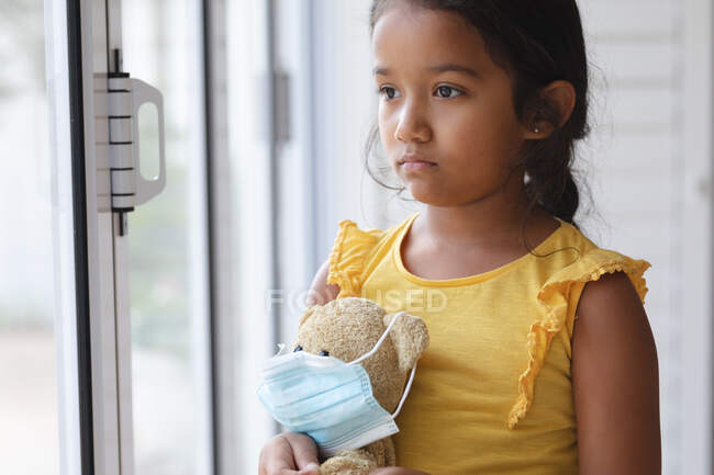 Sad hispanic girl standing at window looking out of window holding teddy bear in face mask. at home in isolation during quarantine lockdown. — Stock Photo