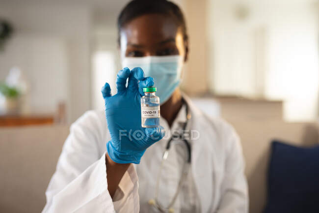 African american female doctor in face mask and gloves holding vial of covid 19 vaccine. medical and healthcare services during coronavirus covid 19 pandemic. — Stock Photo