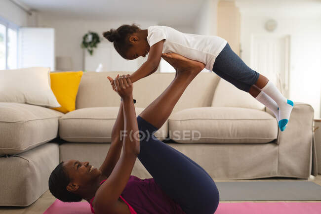 Sonriente madre afroamericana haciendo ejercicio, acostada boca arriba, equilibrando a la hija en los pies en la sala de estar. familia pasar tiempo juntos en casa. - foto de stock