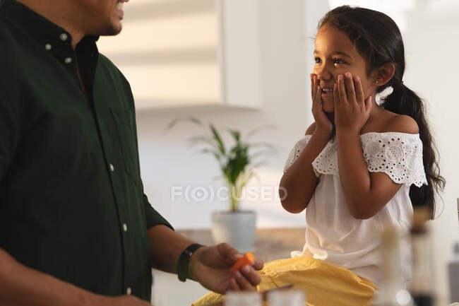 Happy hispanic daughter sitting on counter making face to father preparing vegetables in kitchen. at home in isolation during quarantine lockdown. — Stock Photo
