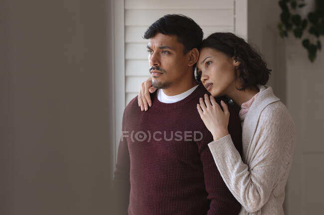 Thoughtful hispanic couple standing at window looking out and embracing. spending free time together at home. — Stock Photo
