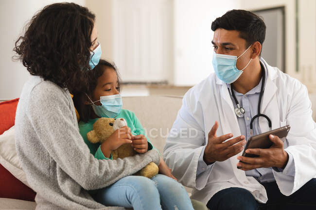 Médico masculino hispano hablando en el sofá en casa de la madre y la hija paciente, todos con máscaras faciales. servicios médicos y sanitarios durante la pandemia de coronavirus covid 19. - foto de stock