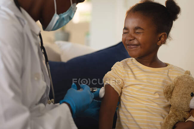 African american female doctor in face mask giving covid vaccination to scared girl patient at home. medical and healthcare services during coronavirus covid 19 pandemic. — Stock Photo