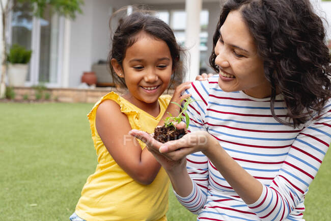 Sonriendo madre hispana y jardinería hija, madre sosteniendo plántulas en la mano. familia pasar tiempo juntos en casa. - foto de stock