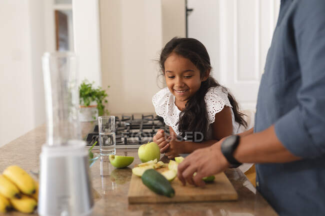 Sonriente hija hispana apoyada en el mostrador viendo a padre preparar verduras en la cocina. en casa en aislamiento durante el bloqueo de cuarentena. - foto de stock