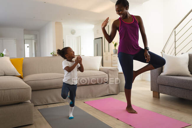 Счастливые африканские американские мать и дочь практикуют йогу, стоя на одной ноге в гостиной. Семья проводит время вместе дома. — стоковое фото