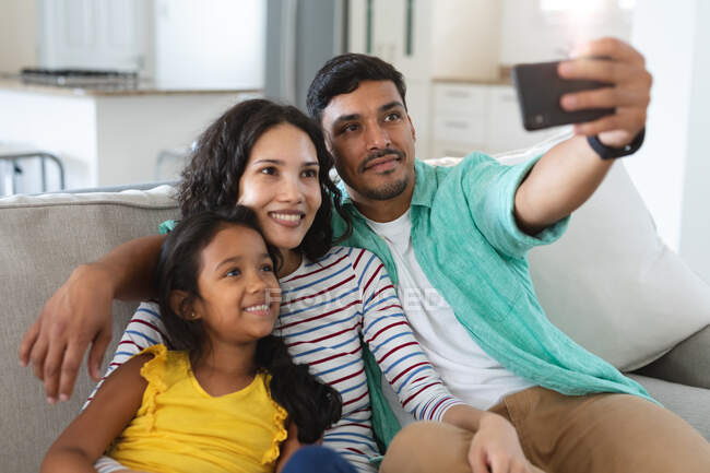 Посміхаючись своїй іспанській матері, батько і дочка сидять на дивані, беручи селфі разом. сім'я проводить час разом вдома . — стокове фото