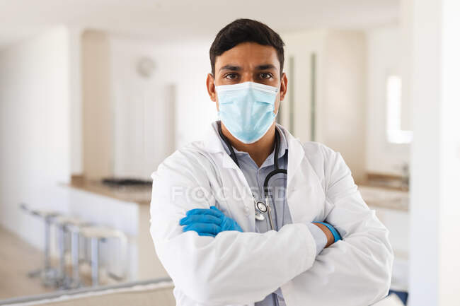 Retrato de un médico hispano con máscara facial de pie con los brazos cruzados mirando a la cámara. servicios médicos y sanitarios durante la pandemia de coronavirus covid 19. - foto de stock