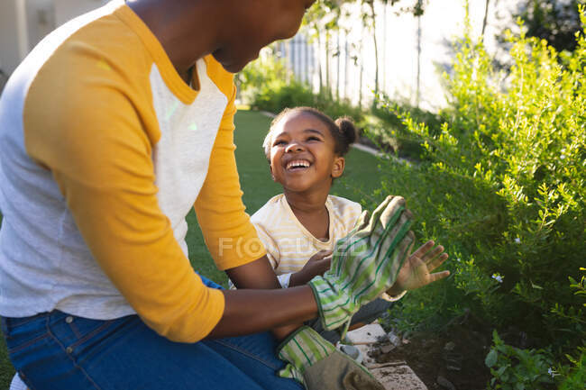 Sorrindo afro-americana mãe e filha ajoelhados cuidando de plantas em jardim ensolarado. família passar tempo juntos em casa. — Fotografia de Stock