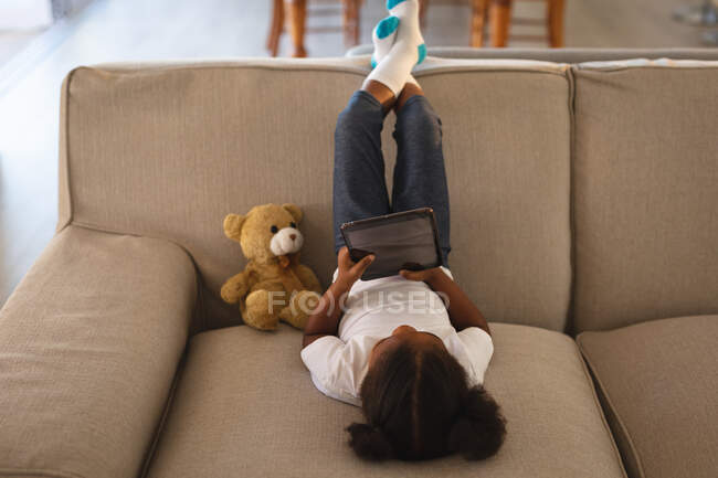Chica afroamericana feliz sentado boca abajo en el sofá usando tableta, con oso de peluche. pasar tiempo libre en casa. - foto de stock