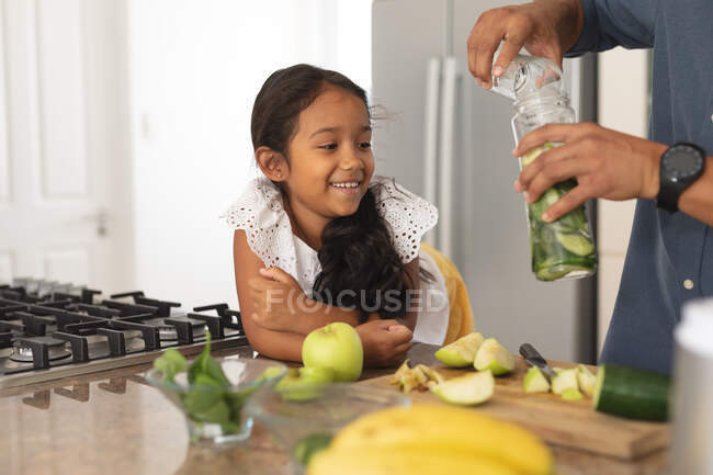 Улыбающаяся латиноамериканская дочь, опираясь на прилавок, наблюдая за тем, как отец готовит напиток на кухне. в доме в изоляции во время карантинной изоляции. — стоковое фото
