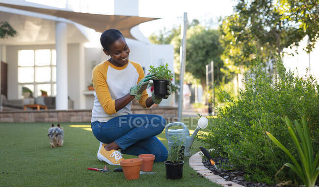 Lächelnde Afroamerikanerin bei der Gartenarbeit, kniend im Garten, Haus im Hintergrund. Freizeit zu Hause verbringen. — Stockfoto