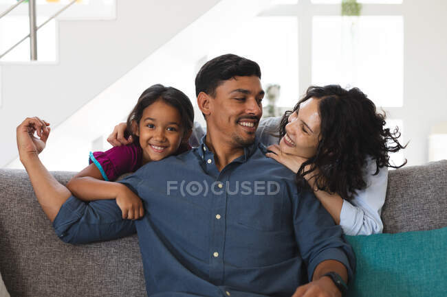 Sonriente padre hispano sentado en el sofá con esposa e hija detrás, abrazándolo. en casa en aislamiento durante el bloqueo de cuarentena. - foto de stock