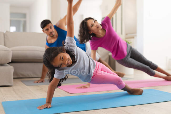 Fille hispanique souriante et parents pratiquant le yoga s'étendant dans le salon. à domicile en isolement pendant le confinement en quarantaine. — Photo de stock