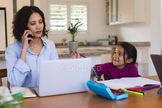 Sonriente hija hispana en la cocina haciendo trabajo escolar con la madre usando smartphone y laptop. familia pasar tiempo juntos en casa. - foto de stock