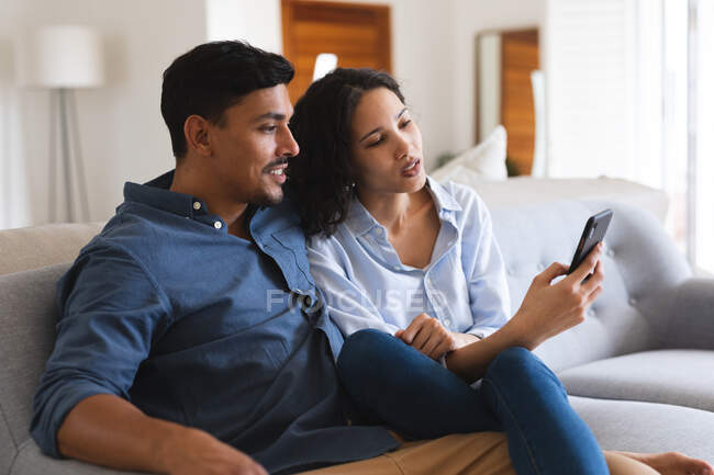 Счастливая испаноязычная пара, сидящая на диване в гостиной со смартфоном. проводить время вместе дома. — стоковое фото