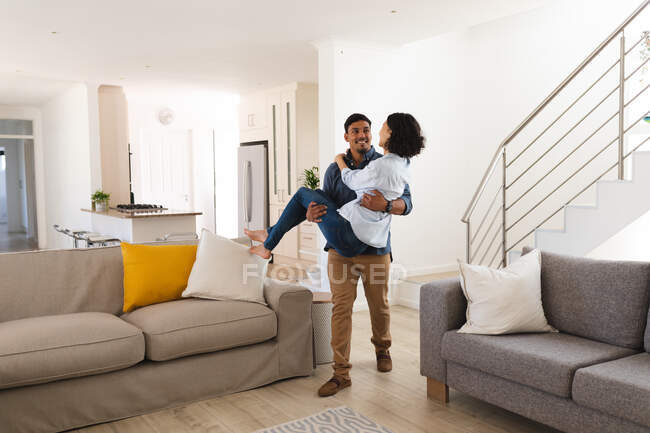 Glückliches hispanisches Paar, das im Wohnzimmer spaziert, Ehemann trägt Frau. Zuhause in Isolation während der Quarantäne. — Stockfoto