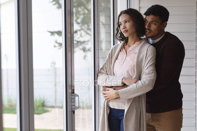 Задумчивая испаноязычная пара, стоящая у окна и обнимающая. проводить свободное время вместе дома. — стоковое фото