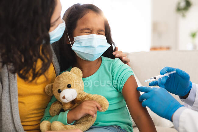 Doctor dando la vacuna covid a la paciente hispana sentada con la madre usando máscaras faciales. servicios médicos y sanitarios durante la pandemia de coronavirus covid 19. - foto de stock