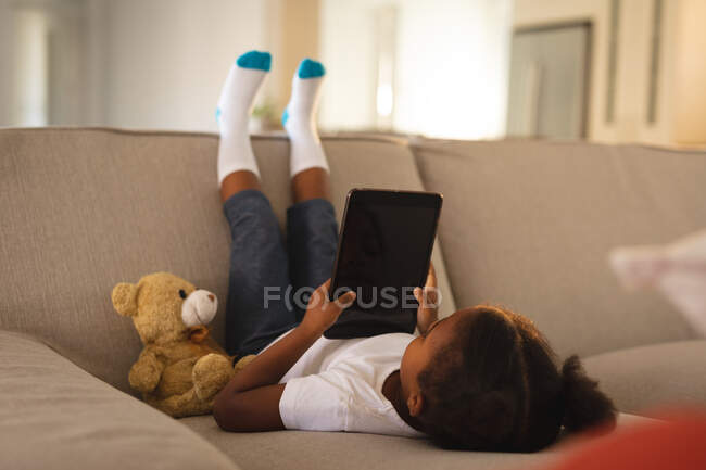 Menina americana africana feliz de cabeça para baixo no sofá usando tablet, espaço de cópia na tela, com ursinho de pelúcia. tempo livre em casa. — Fotografia de Stock
