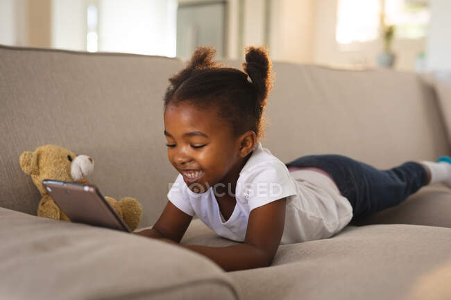 Felice ragazza afroamericana sdraiata sul divano con orsacchiotto, utilizzando tablet e sorridente. trascorrere del tempo libero a casa. — Foto stock