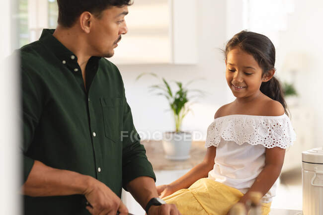 Feliz hija hispana y padre preparando comida en la cocina, hija sentada en el mostrador. en casa en aislamiento durante el bloqueo de cuarentena. - foto de stock