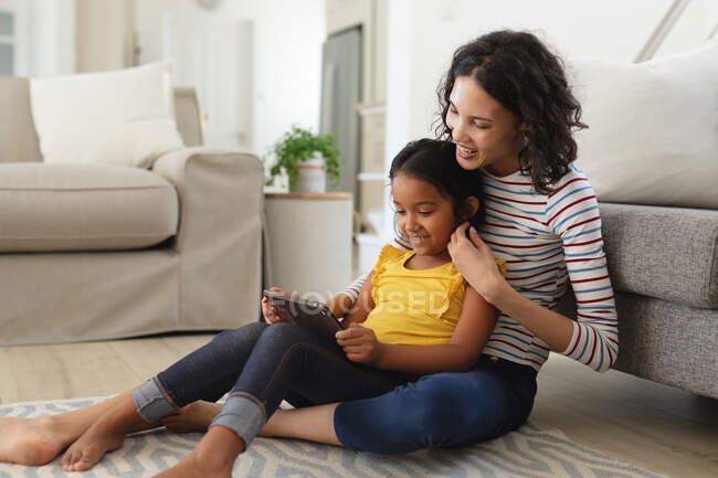 Улыбающиеся латиноамериканская мать и дочь сидят на полу гостиной с помощью планшета вместе. Семья проводит время вместе дома. — стоковое фото