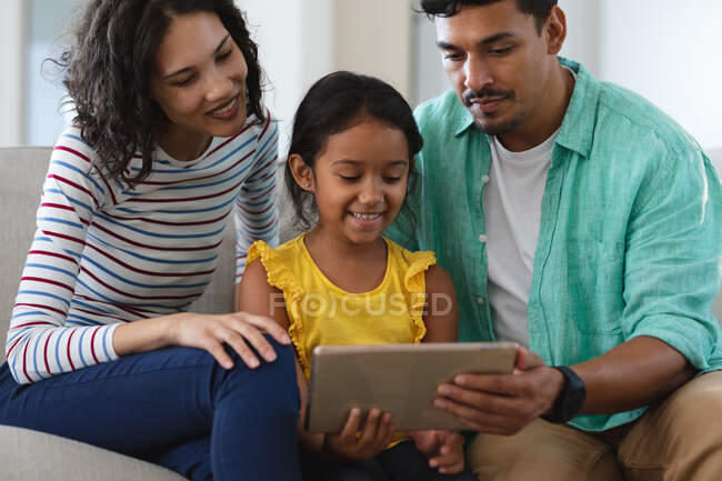 Sorrindo mãe hispânica, pai e filha sentados no sofá usando tablet juntos. família passar tempo juntos em casa. — Fotografia de Stock