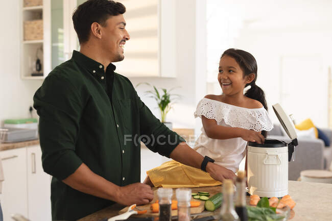 Улыбающиеся латиноамериканка и отец готовят овощи на кухне, дочь сидит на прилавке. в доме в изоляции во время карантинной изоляции. — стоковое фото