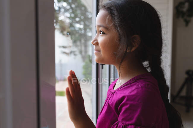 Glückliches hispanisches Mädchen, das mit der Hand auf Glas am Fenster steht, hinausblickt und lächelt. Freizeit zu Hause. — Stockfoto