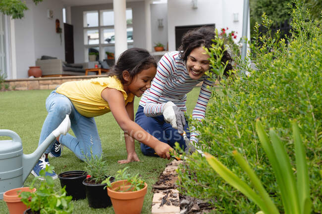 Lächelnde hispanische Mutter und Tochter gärtnern, knien und pflanzen im Blumenbeet. Familie verbringt Zeit zu Hause. — Stockfoto