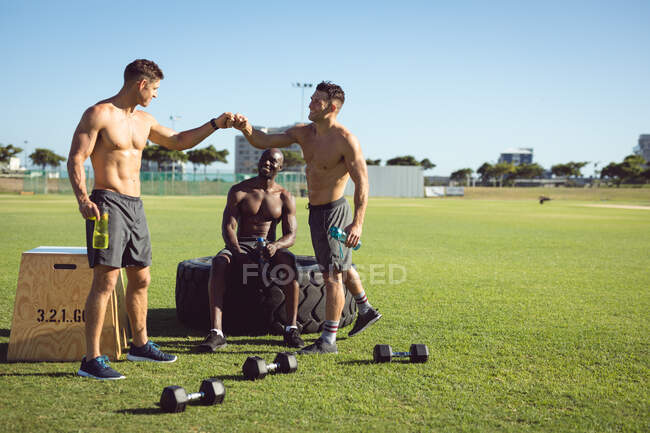 Diverso grupo de hombres felices sin camisa ejercitándose al aire libre, tomando un descanso hablando y golpeando puños. estilo de vida activo saludable, entrenamiento cruzado para fitness. - foto de stock
