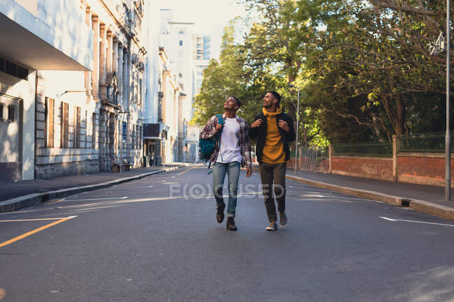 Dos felices amigos varones de raza mixta llevando mochilas caminando por la calle de la ciudad. vacaciones de mochilero, escapada a la ciudad. - foto de stock