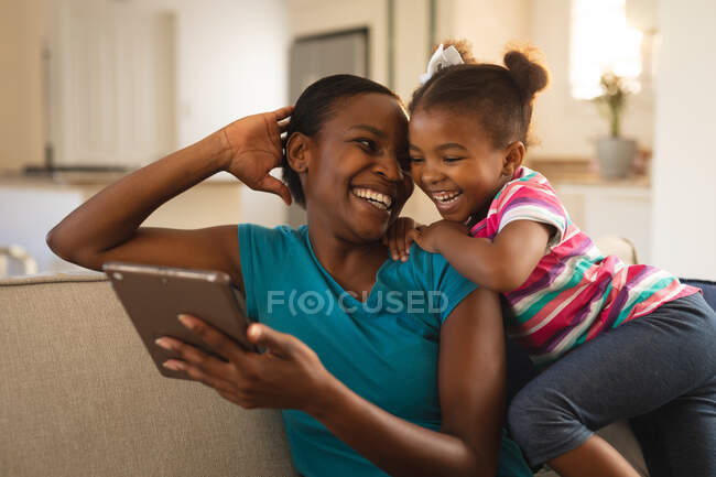 Mãe e filha afro-americana feliz relaxando no sofá olhando para tablet e rindo juntos. família passar tempo juntos em casa. — Fotografia de Stock