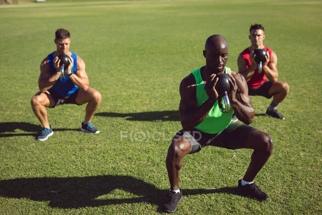 Разнообразная группа мускулистых мужчин, тренирующихся с колокольчиками чайника на открытом воздухе. здоровый активный образ жизни, перекрестные тренировки для фитнеса. — стоковое фото