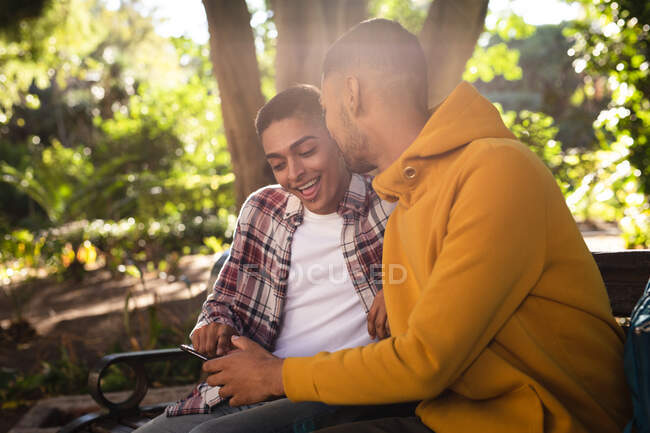 Dos amigos varones de raza mixta felices sentados en el banco del parque usando un teléfono inteligente. vacaciones de mochilero, escapada a la ciudad. - foto de stock