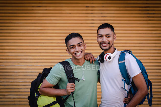 Retrato de dos amigos varones de raza mixta felices con mochilas en la calle de la ciudad, sonriendo y abrazando. vacaciones de mochilero, escapada a la ciudad. - foto de stock