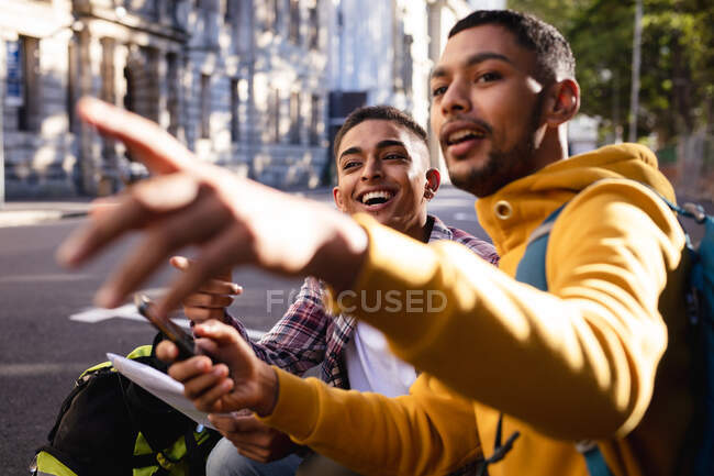 Dos amigos varones de raza mixta sonrientes sentados en la calle, usando un teléfono inteligente y apuntando hacia la dirección. vacaciones de mochilero, escapada a la ciudad. - foto de stock