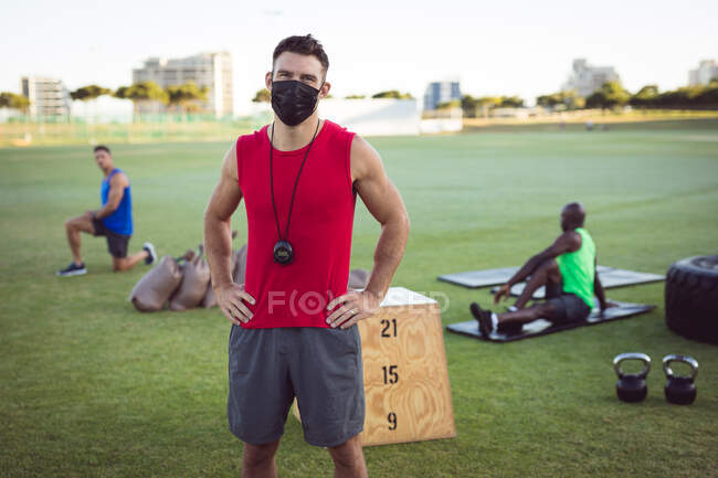 Portrait of caucasian male fitness trainer wearing face mask, with colleagues exercising outdoors. — Stock Photo
