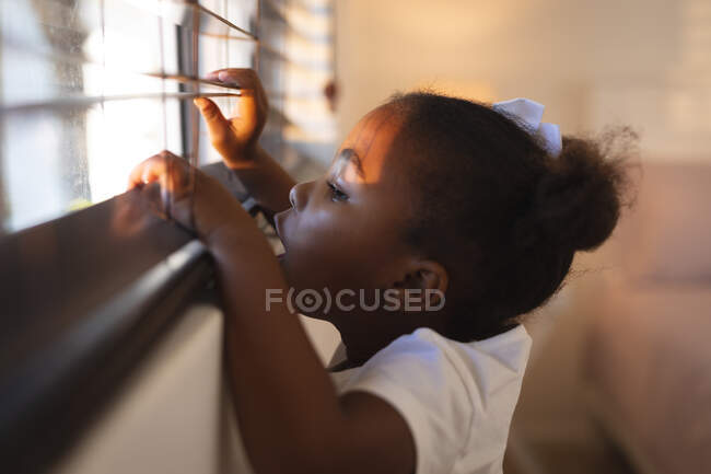 Curiosa ragazza afroamericana in piedi e sbirciando attraverso le tende delle finestre in una giornata di sole. trascorrere del tempo libero a casa. — Foto stock